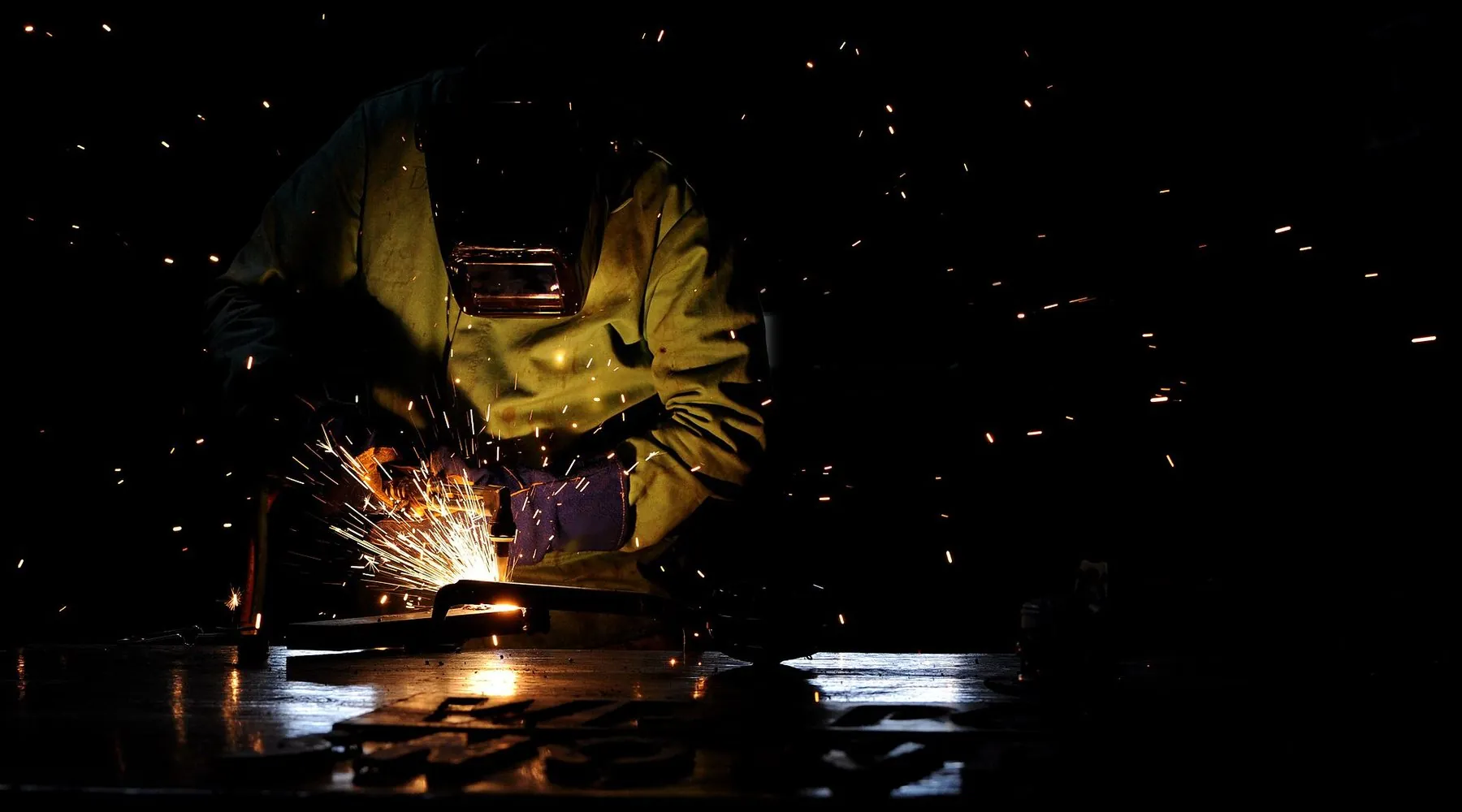 Welder in a protective jacket and helmet cutting steel, sparks flying in a dark workshop