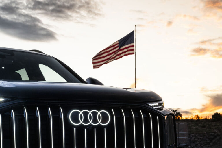 Front grille and illuminated four-ring logo of an Audi Q9 SUV with a U.S. flag flying behind it at sunset.