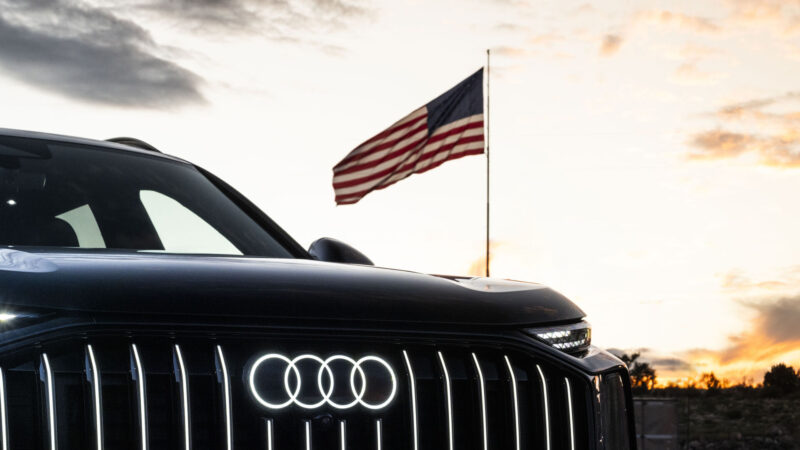 Front grille and illuminated four-ring logo of an Audi Q9 SUV with a U.S. flag flying behind it at sunset.